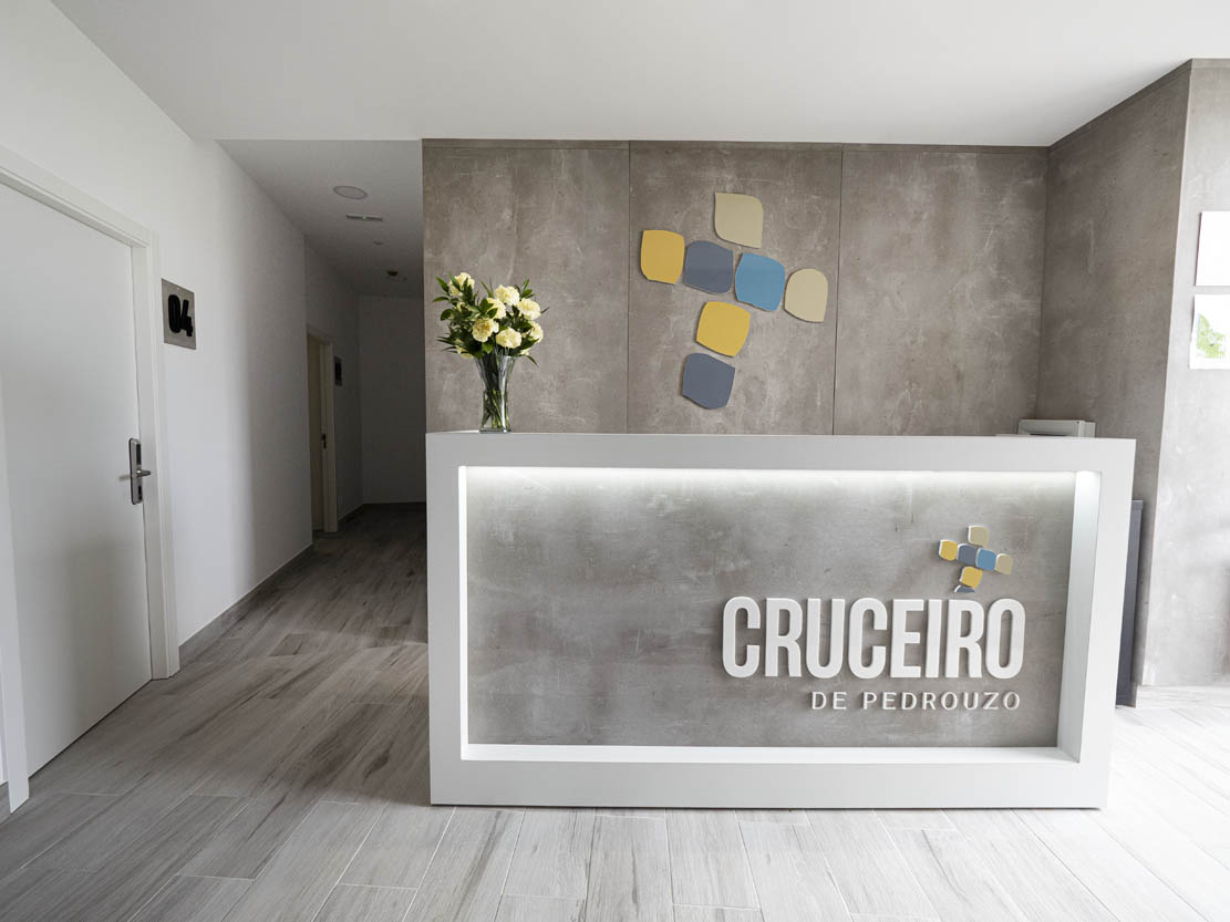 Recepción del albergue Mostrador de recepción moderno con iluminación frontal y el rótulo de “Cruceiro de Pedrouzo”, acompañado de un jarrón con flores y un logotipo de figuras de colores en la pared.
